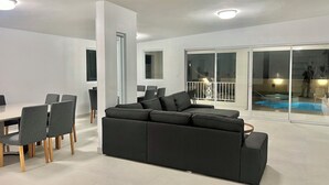 A modern living room with a sectional sofa, dining table with chairs, and a glass door leading to a balcony with a pool.