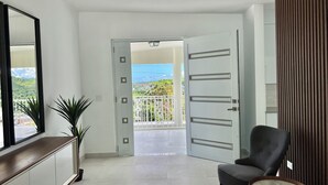 A modern interior with a white tiled floor, a dark armchair, a small round table, a potted plant, and a view of a balcony with a railing and greenery.