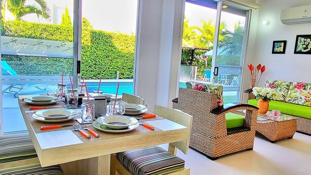 Un comedor con una mesa dispuesta para cuatro personas, un sofá de mimbre y vista a una piscina.