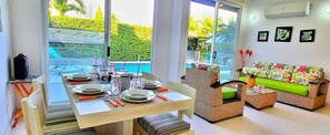 A dining area with a table set for four, a wicker sofa, and a view of a swimming pool. - Encantadora Casa Piscina Privada AA Wifi (Ricaurte)
