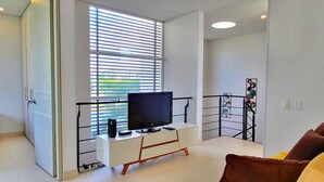 Una sala de estar moderna con un televisor sobre un mueble blanco, un balcón con barandilla y un sofá con cojines.