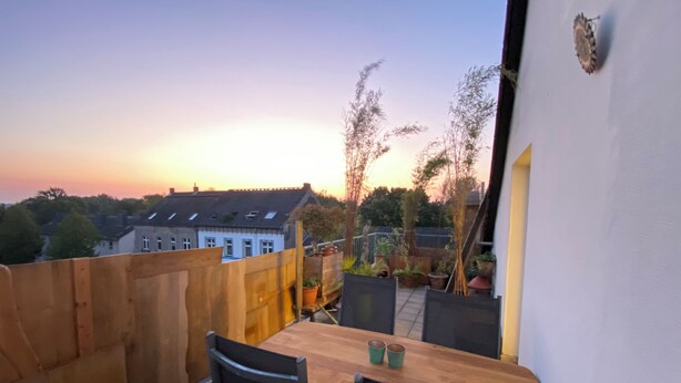 Ein Balkon mit Holztisch und -stühlen, Blumentöpfen und Blick auf Wohnhäuser bei Sonnenuntergang.