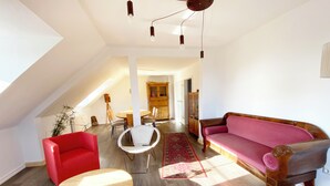 Ein großzügiger Wohnraum mit einem roten Sessel, einem Holztisch, einem rosa Sofa und einer Essecke mit rundem Tisch und Stühlen.