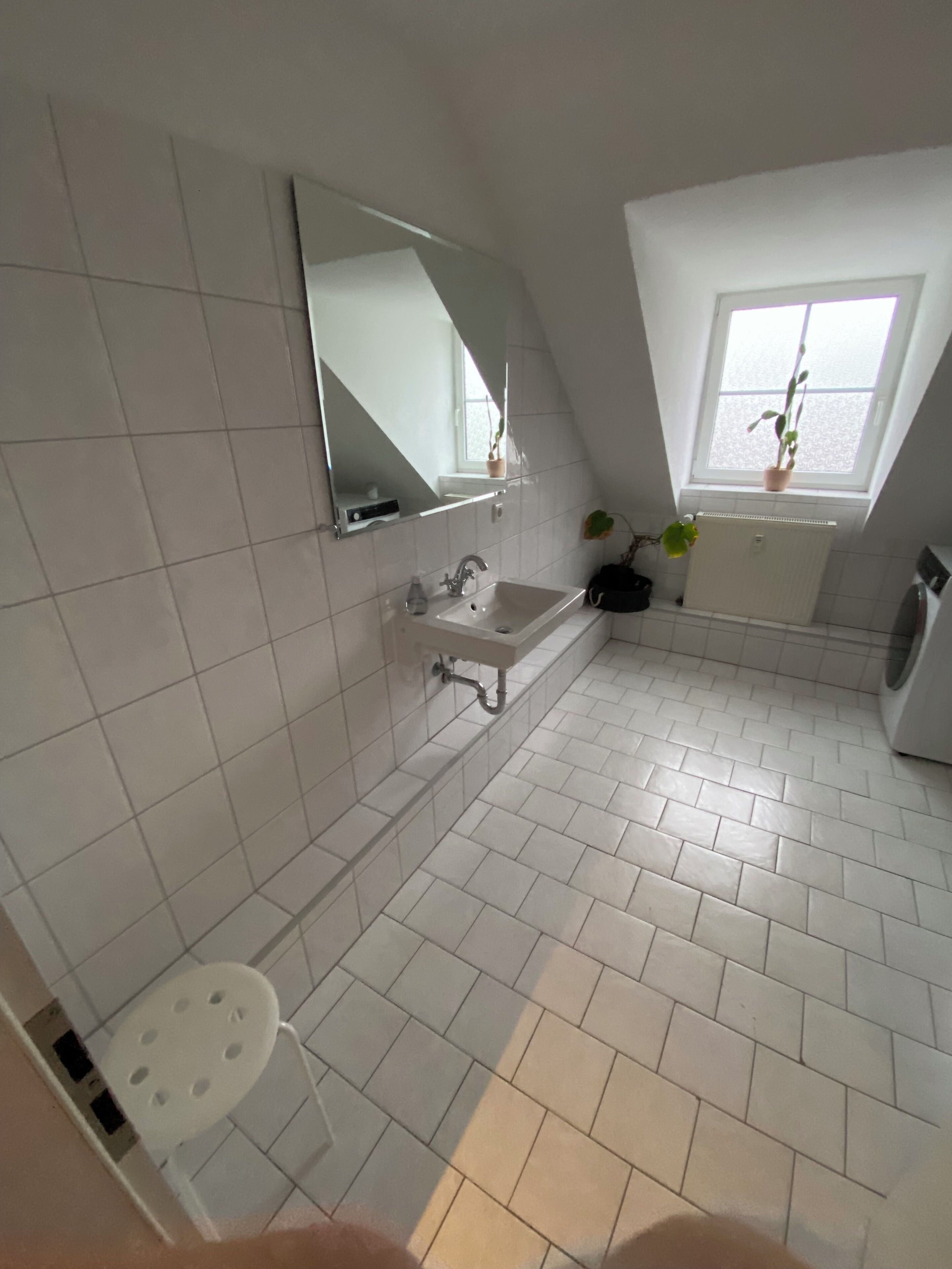 A modern bathroom with white tiles, a sink, a mirror, a window with a plant, and a washing machine.