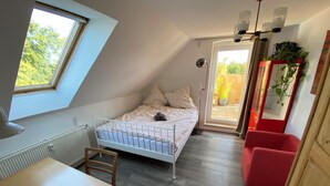 Ein kleiner, gemütlicher Raum mit schrägem Dach, einem Einzelbett, einem Holztisch und einem roten Stuhl.