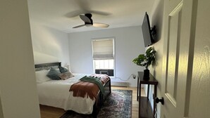 Une chambre à coucher avec un lit, un ventilateur de plafond, une fenêtre, un téléviseur fixé au mur et une plante en pot.