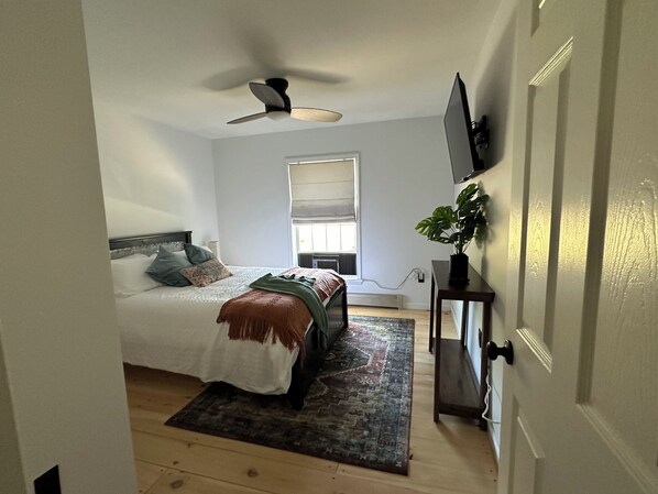 Ein Schlafzimmer mit Bett, Deckenventilator, Fenster, an der Wand montiertem Fernseher und einer Zimmerpflanze.