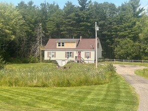Une maison à deux étages avec une porte rouge et des volets marron, entourée d’une clôture en bois et bordée d’un chemin de gravier.