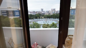 En balkong med utsikt över en stadsbild, inklusive en rund fontän och olika byggnader. Det står två stolar och ett bord med frukt på balkongen, och en bok ligger på bordet.