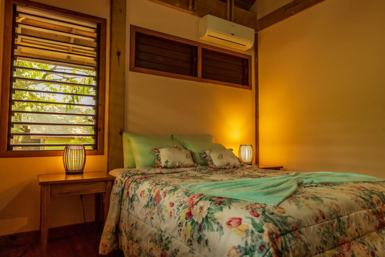 A bedroom with a bed, a nightstand, a window with blinds, and an air conditioning unit.