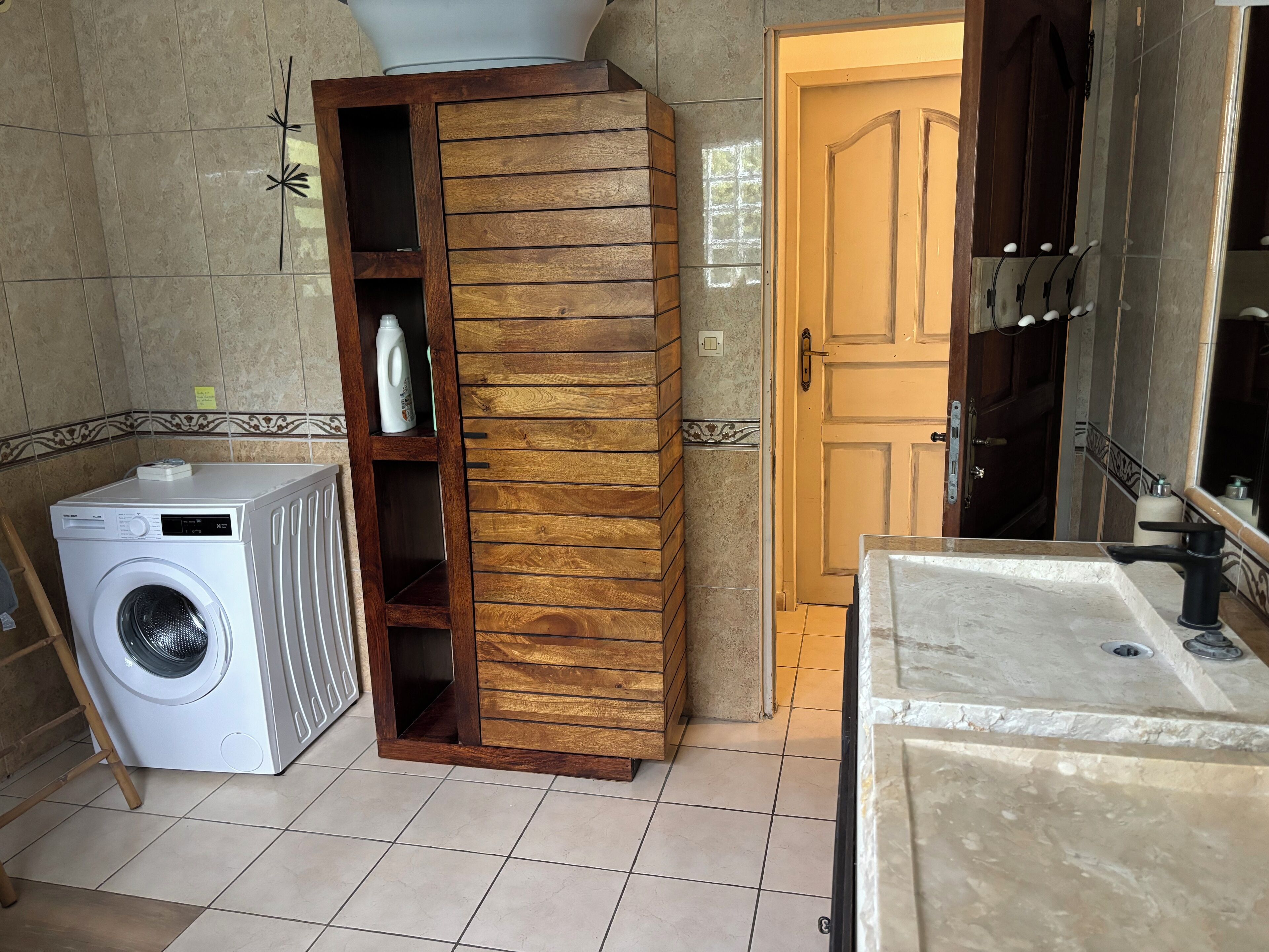 Un lavadero pequeño con lavadora, un armario de madera con estantes, y un baño con lavabo y espejo.