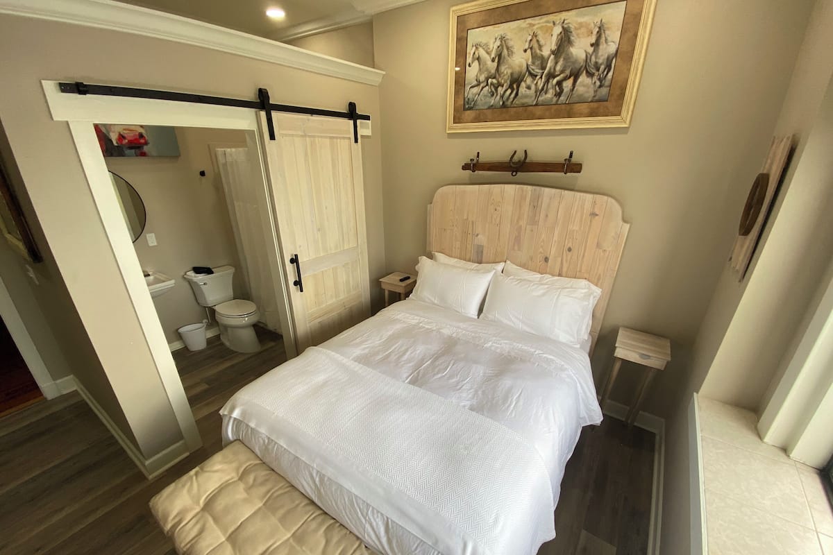 A bedroom with a bed, a bench, a wooden headboard, a painting, and a bathroom visible through an open door.