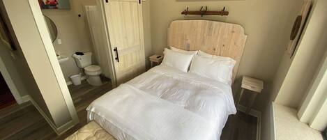 Ein Schlafzimmer mit Bett, einer Sitzbank, einem Holzkopfteil, einem Gemälde und einem durch eine offene Tür einsehbaren Badezimmer.