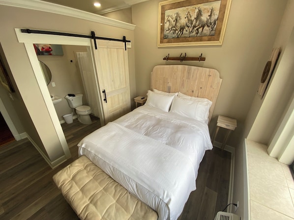A bedroom with a bed, a bench, a wooden headboard, a painting, and a bathroom visible through an open door. - The Roanoke Hotel (Sebring)
