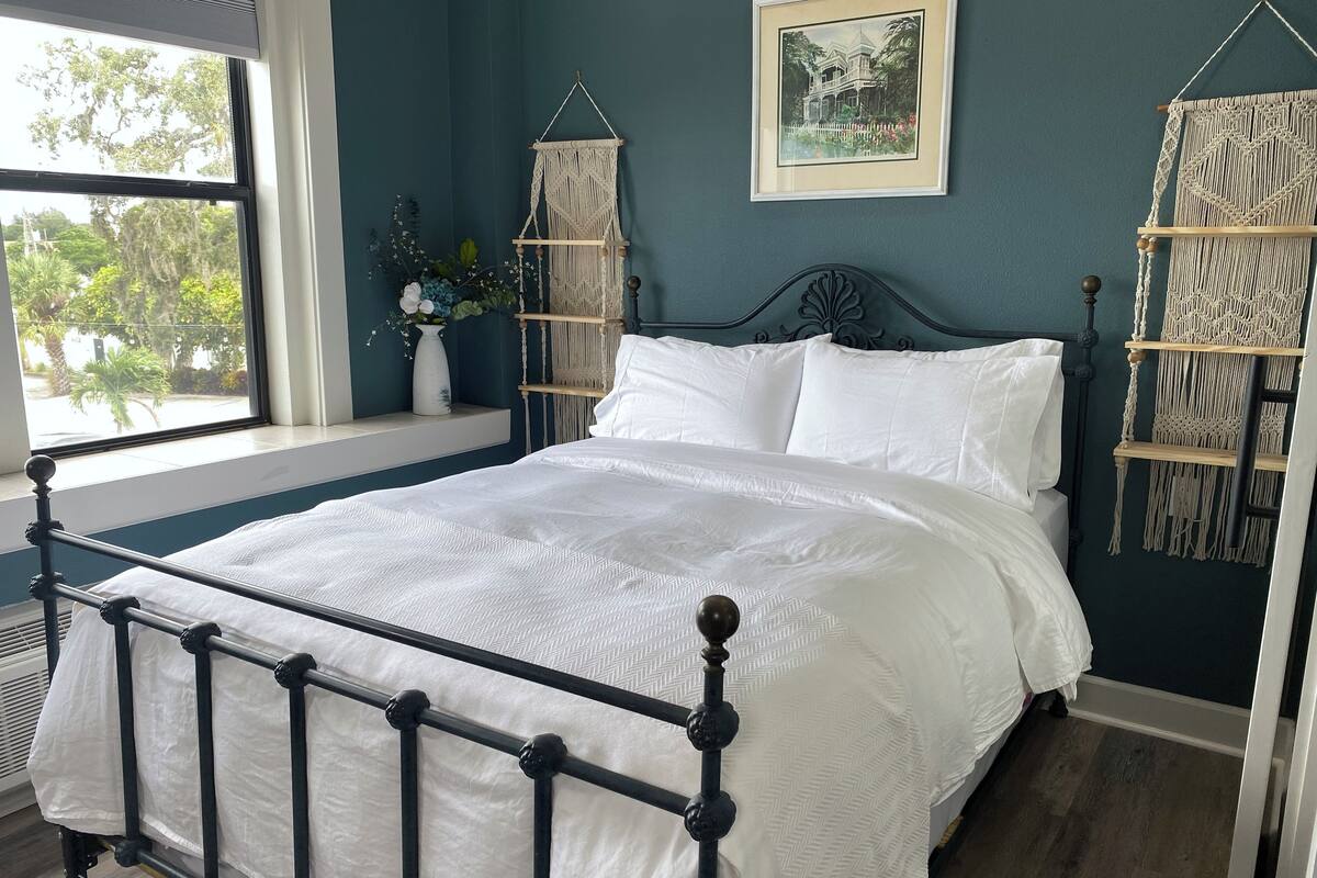 A bedroom with a bed, a window, a macrame wall hanging, and a framed picture.
