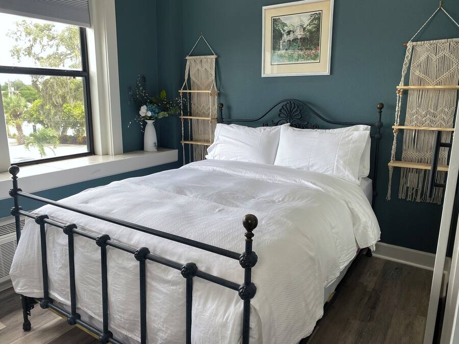 Ein Schlafzimmer mit Bett, Fenster, Wandbehagung aus Makramee und einem gerahmten Bild.