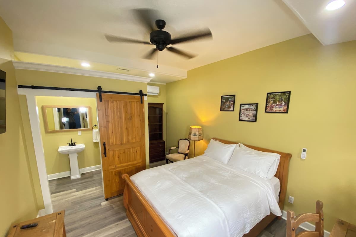 A bedroom with a bed, a wooden door, a mirror, a bedside table, a lamp, and a wall-mounted air conditioner.