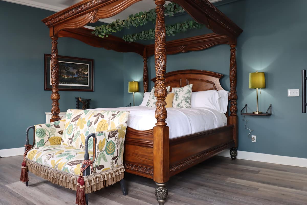 A four-poster canopy bed with a floral-patterned chair, a framed picture on the wall, and two wall-mounted lamps.