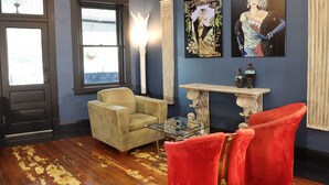 A room with a dark blue wall, a wicker armchair, a red velvet armchair, a glass coffee table, and two paintings. - The Roanoke Hotel (Sebring)
