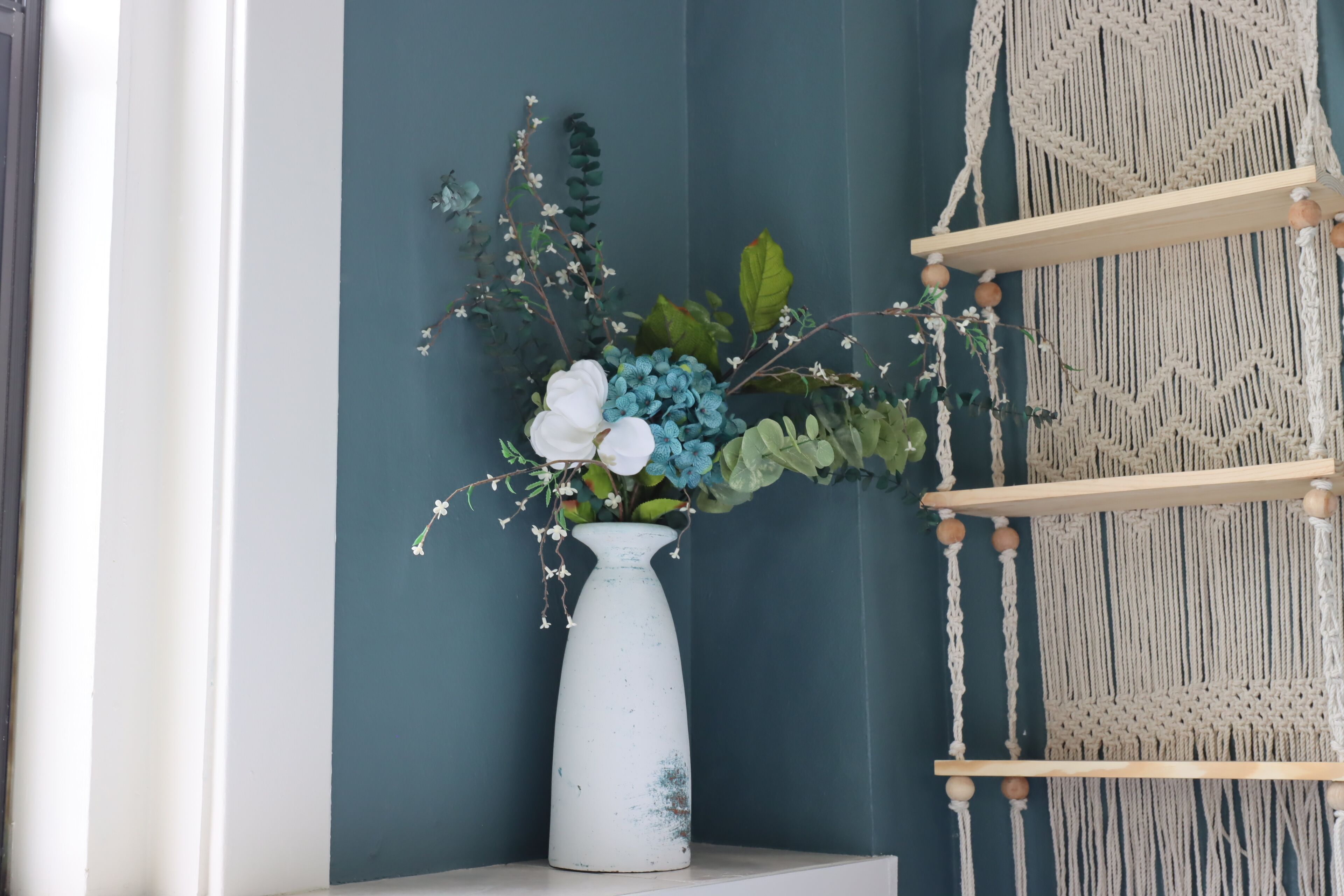 A white vase with flowers on a white shelf against a teal wall, with a macrame wall hanging to the right.