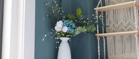 Un jarrón blanco con flores sobre una estantería blanca frente a una pared color verde azulado, con un colgador de macramé a la derecha.