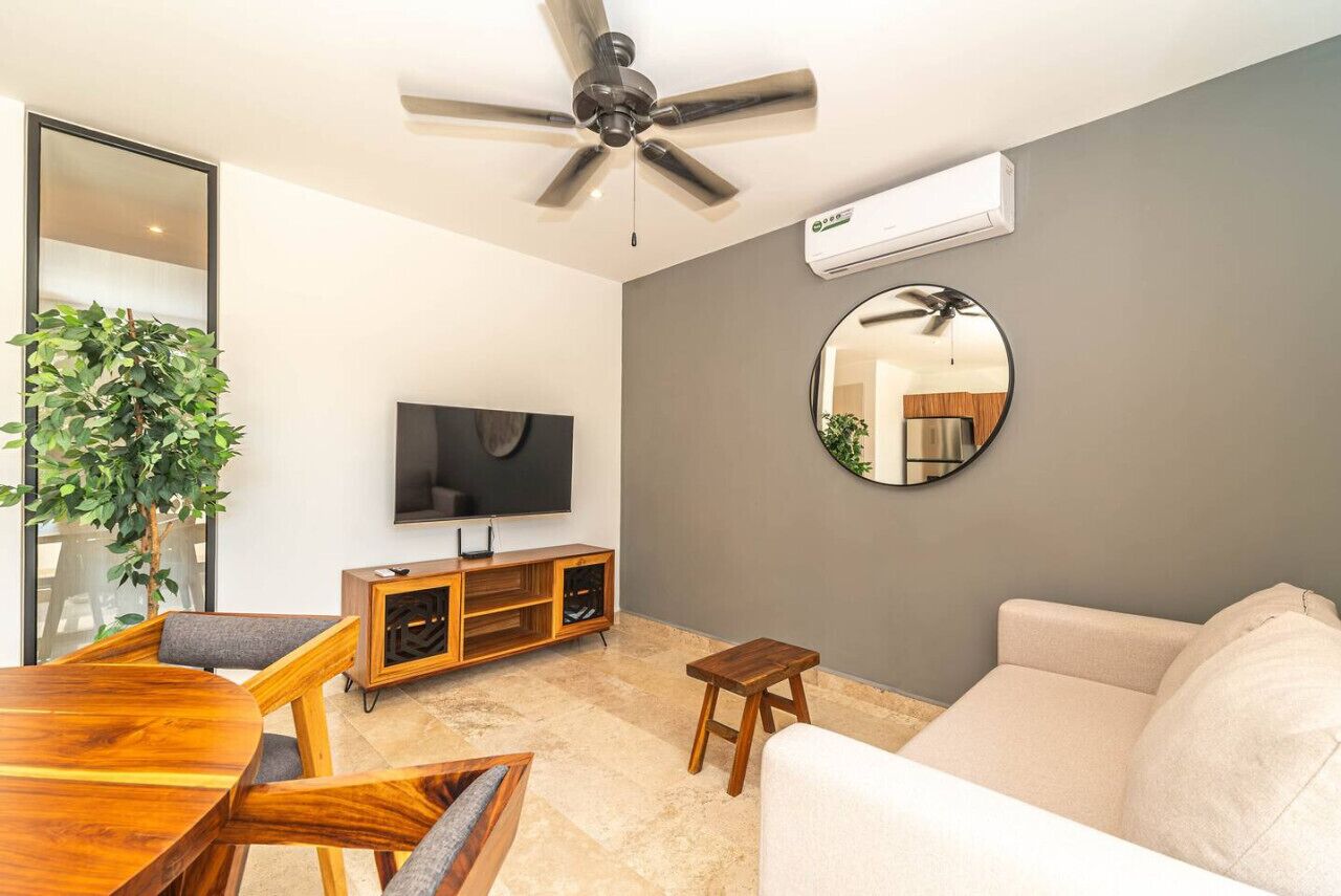 Una sala de estar moderna con ventilador de techo, televisor de pantalla plana, espejo redondo, mueble bajo para televisión de madera, sofá color beige y una mesa de comedor con sillas.