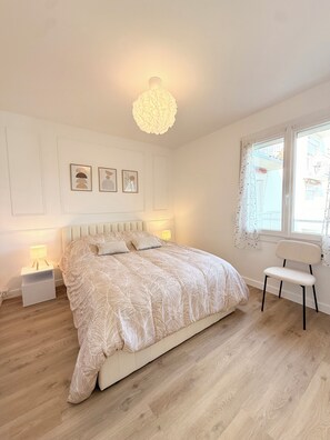 Une chambre à coucher comprenant un lit, des tables de chevet, une chaise, une fenêtre avec des rideaux et des œuvres d’art accrochées au mur.