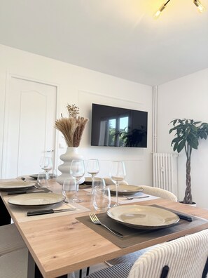 Un coin repas avec une table en bois dressée pour quatre personnes, un vase blanc contenant des plantes séchées, une plante en pot et un téléviseur fixé au mur.