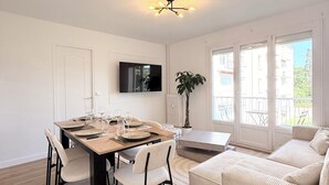 A modern living room with a dining area, a flat-screen TV mounted on the wall, and a large window with a view of buildings.