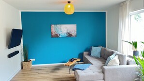 En stue med blå væg, ophængt TV, grå sofa med puder, sofabord i træ og en plante.