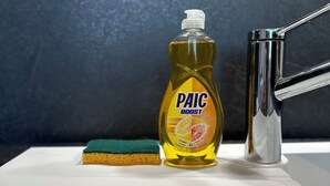 Une bouteille de liquide vaisselle Paic Boost, une éponge verte et un robinet chromé sur un évier blanc.