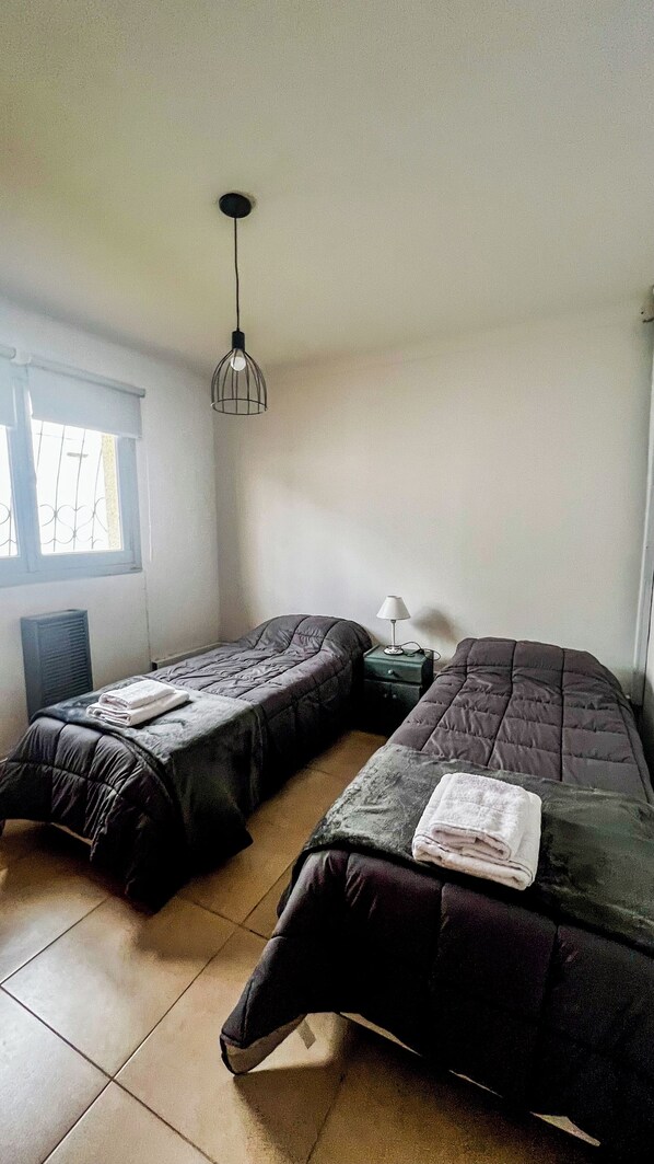 Una habitación con dos camas, un radiador, una ventana con rejas y una lámpara colgante.