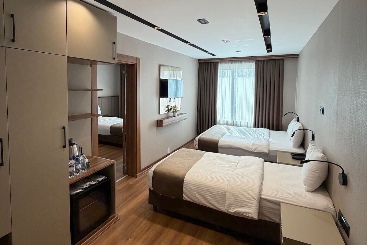 Ein modernes Hotelzimmer mit Holzboden, einem großen Schrank, einem Bett mit Kopfteil, einem Nachttisch und einem Vorhangfenster.
