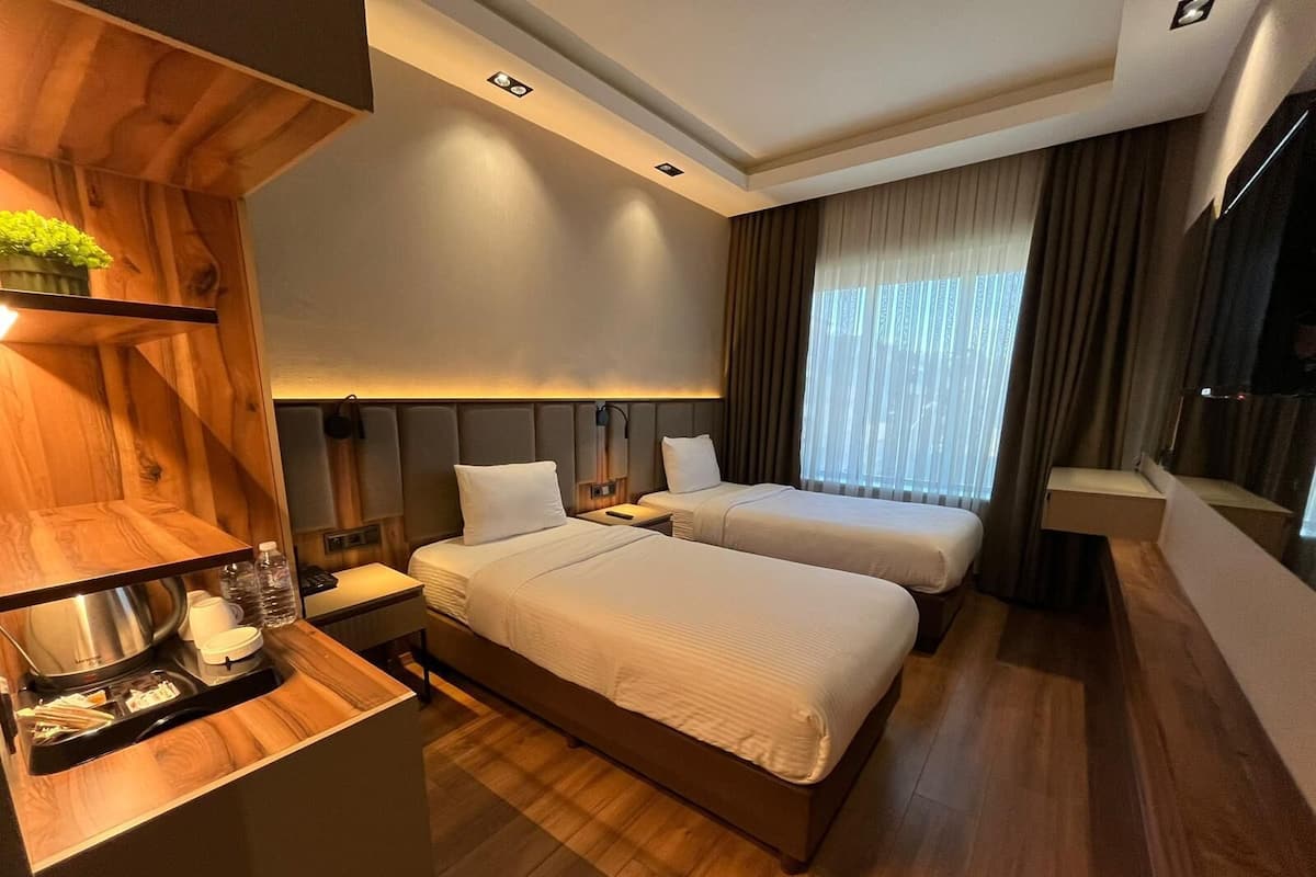 Ein Hotelzimmer mit zwei Betten, einem Holztisch, einer kleinen Küchenzeile und einem großen Fenster mit Vorhängen.