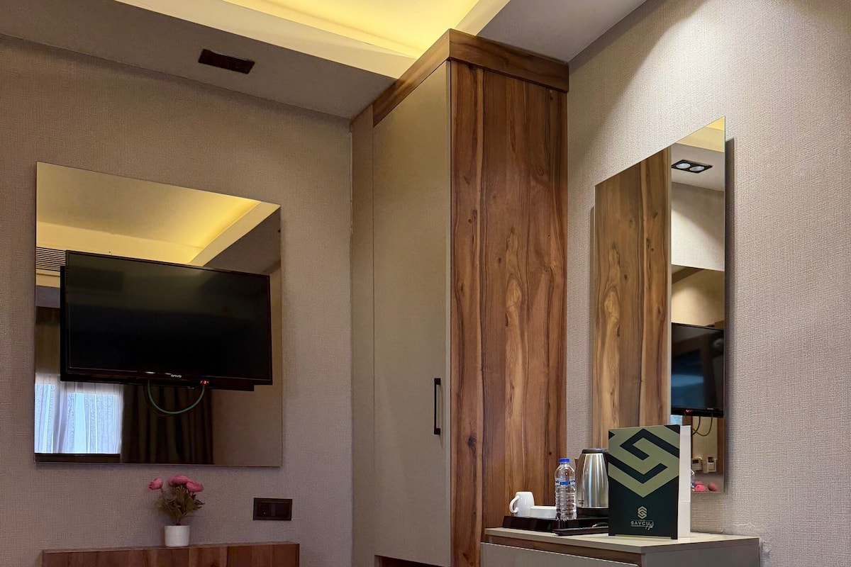 Ein Hotelzimmer mit einem Bett, einem an der Wand befestigten Fernseher, einem Holzschrank, einem Schreibtisch mit Spiegel sowie einem kleinen Tisch mit einer Wasserflasche und einer Kaffeetasse.