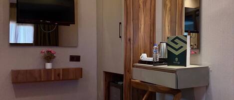 A hotel room with a bed, a TV mounted on the wall, a wooden wardrobe, a desk with a mirror, and a small table with a water bottle and a coffee cup.
