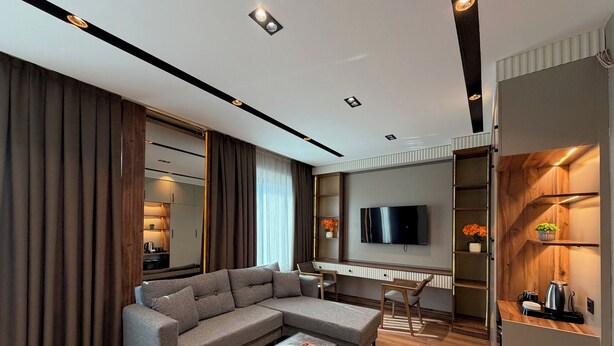 A modern living room with a sectional sofa, a coffee table, a TV mounted on the wall, and a wooden cabinet with shelves. - GRAND SAVCILI DELUX (Gaziantep)