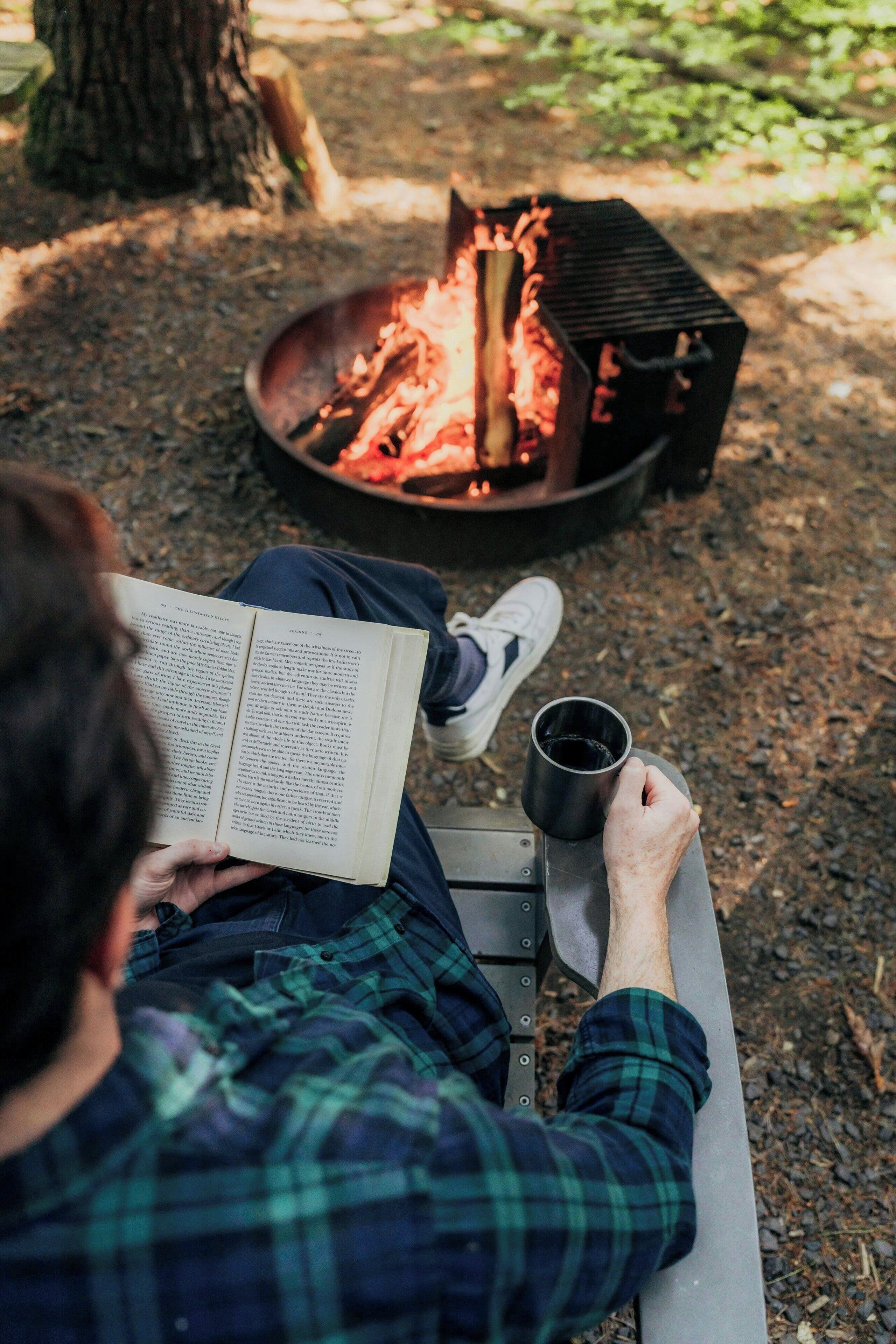 Eine Person sitzt auf einer Bank und liest ein Buch, im Hintergrund brennt ein Lagerfeuer.
