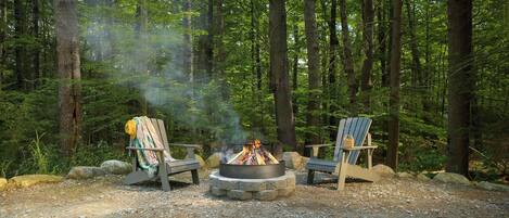 A campfire with two Adirondack chairs, surrounded by a forest.