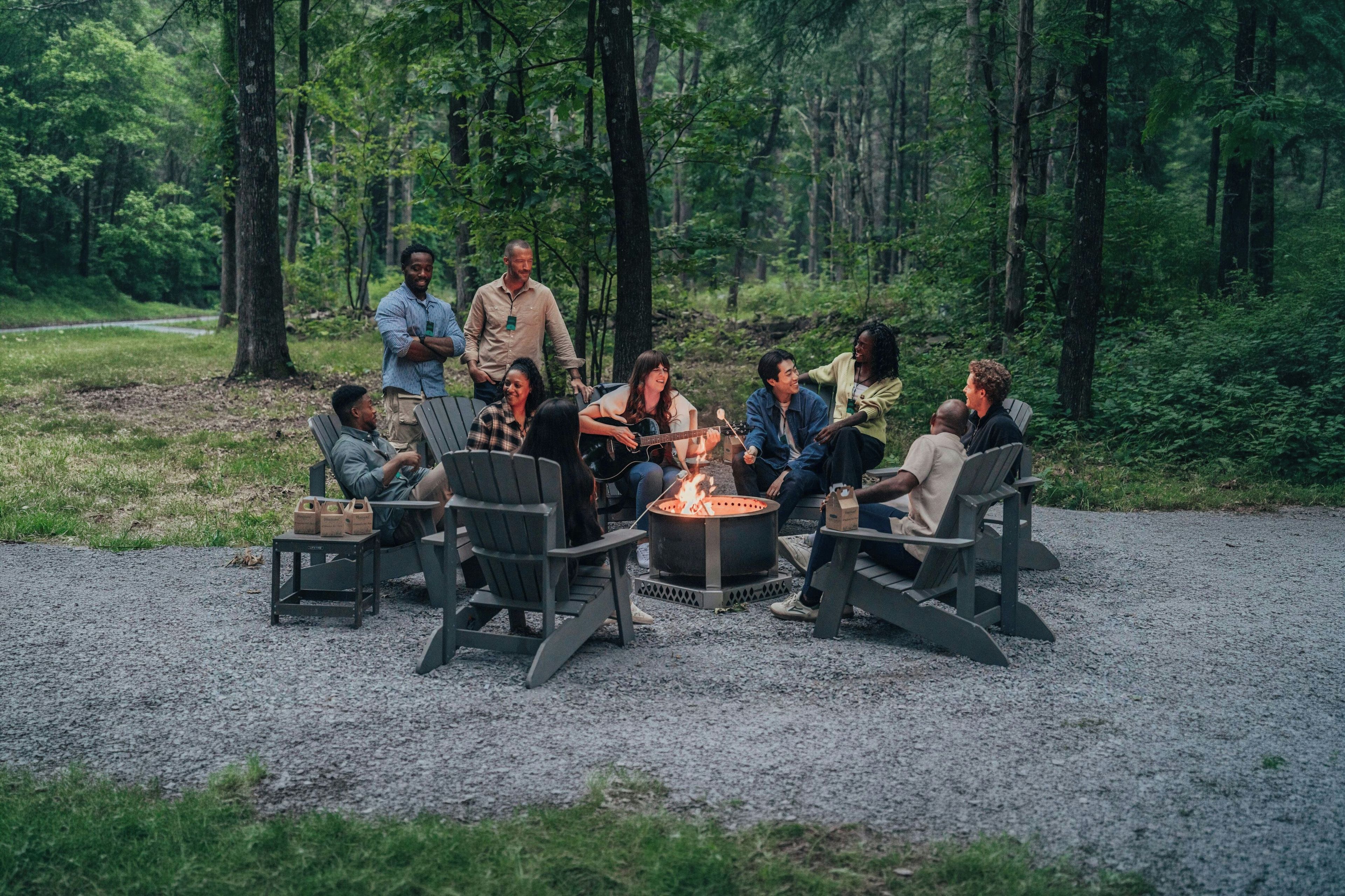 A group of people gathered around a fire pit in a forested area with outdoor seating.