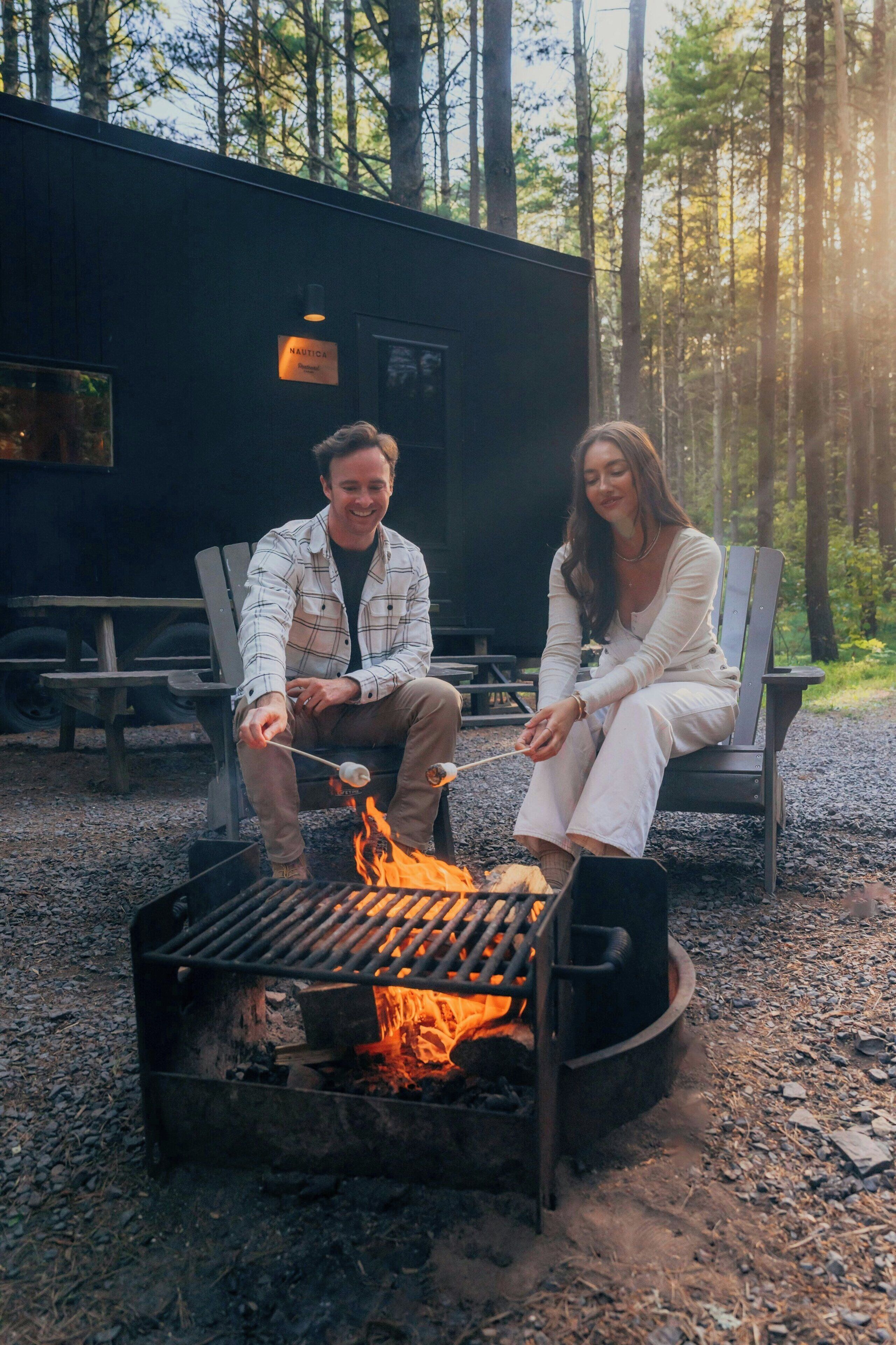 Two people sitting by a campfire in a forested area with a cabin in the background.