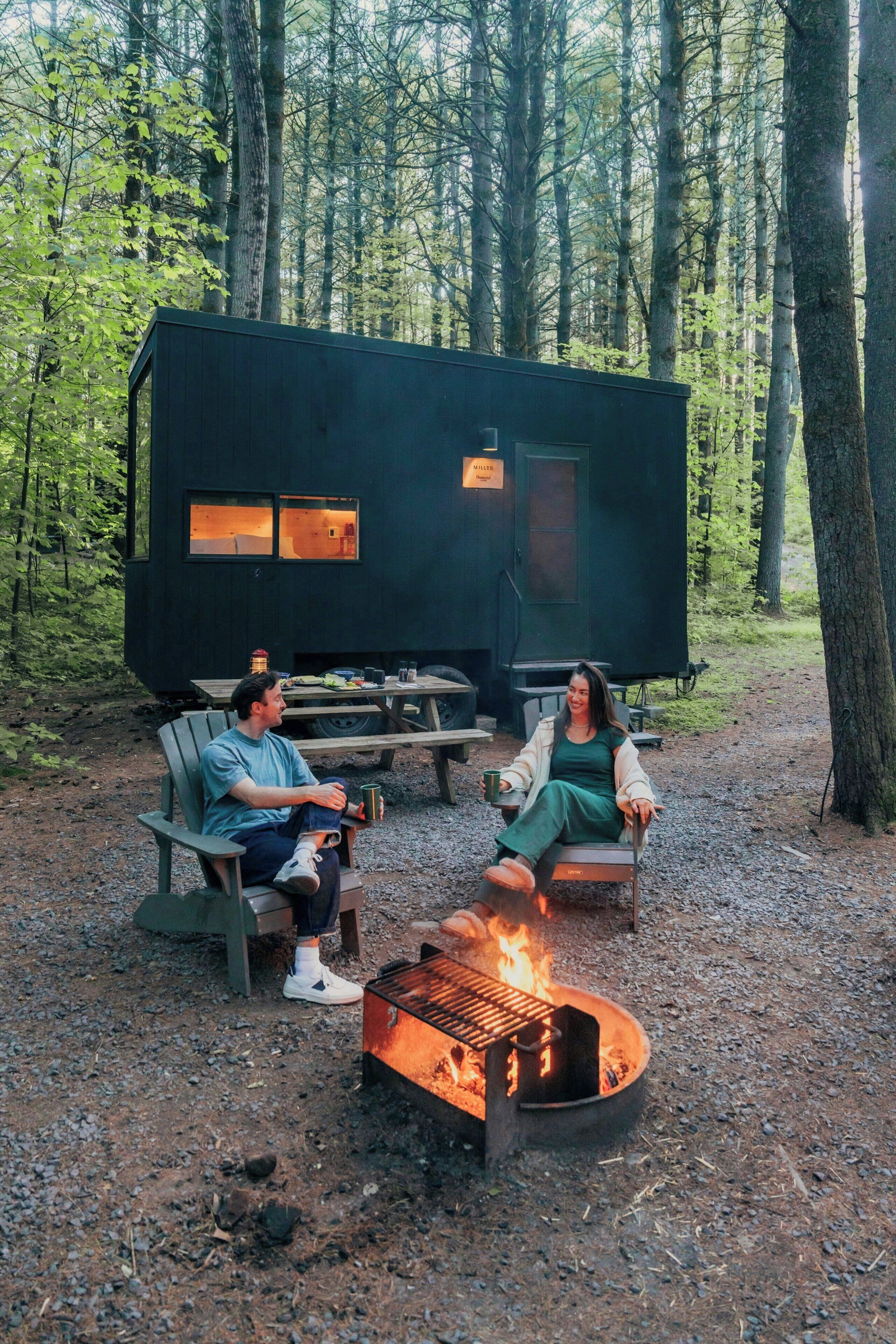 A cabin with a fire pit in the foreground, surrounded by a forest.
