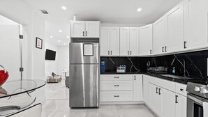 Cocina moderna con armarios blancos, refrigerador de acero inoxidable, encimera de mármol negro y mesa de comedor de vidrio.