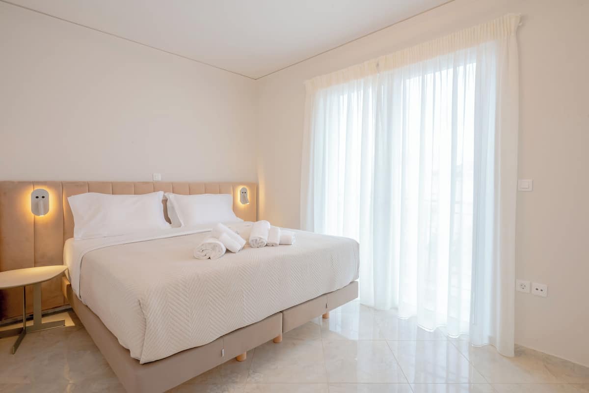 A modern hotel room with a large bed, a bedside table, and a large window with sheer curtains.