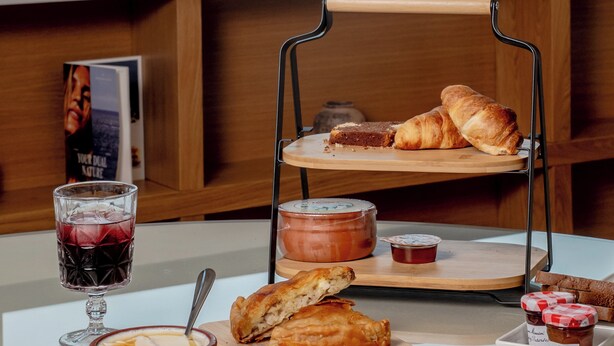 Disposição para o pequeno-almoço com croissants, pão e pastelaria num suporte de madeira, uma taça de sopa e um copo de vinho tinto numa mesa com vista para um edifício histórico ao fundo.