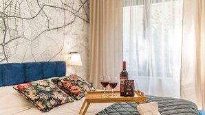 Een hotelkamer met een bed, bloemenkussens, een dienblad met wijnglazen en een fles, en een kaart aan de muur.