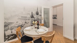 Een eetruimte met een ronde tafel voor vier personen, een vintage stadsgezicht muurschildering op de muur, en een keuken zichtbaar door een open deur.