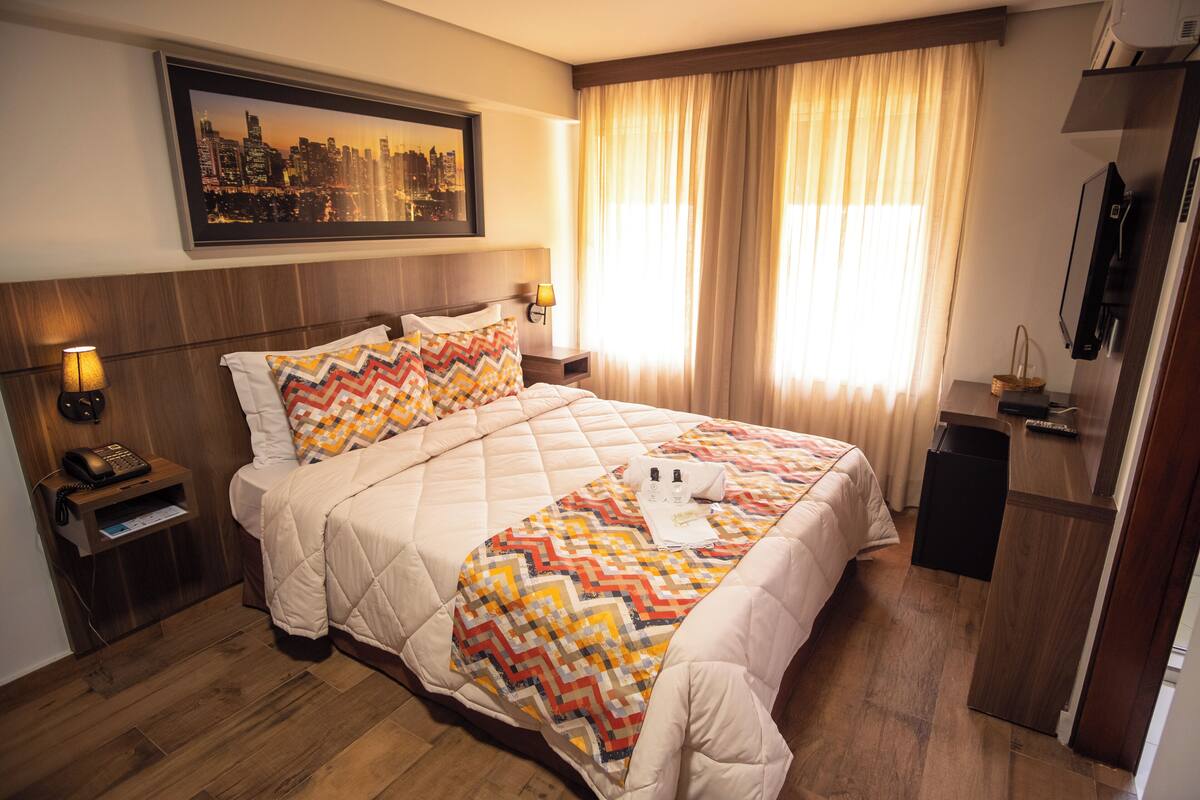 Hotellihuone, jossa on suuri sänky, kaupunkimaisemaa esittävä taulu, työpöytä lampun kanssa ja televisio.