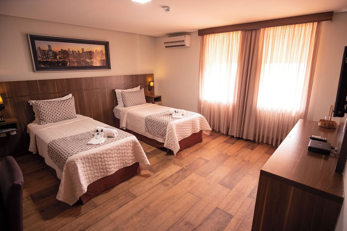 Hotellihuone, jossa on kaksi sänkyä, kaupunkimaisemaa esittävä taulu, ilmastointilaite ja puinen työpöytä, jossa on kaukosäädin.