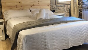 Un dormitorio con pared de piedra, cabecero de madera, cama con ropa blanca y ventana con cortinas.